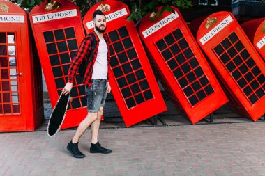 sakallı bir adam, elinde kaykay tutan ve kırmızı bir telefon kulübesiönünde poz veren bir adam