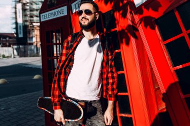 Sakallı şık adam, hipster elinde bir kaykay tutar ve kırmızı bir telefon kulübesi önünde pozlar