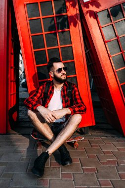 Sakallı şık adam, kaykayile poz veren hipster, şehirde kırmızı bir telefon kulübesinin önünde oturuyor..