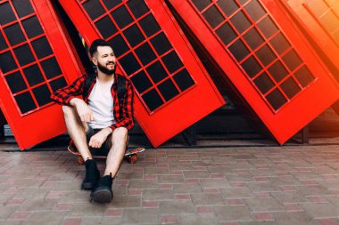 Sakallı şık adam, elinde kaykay tutan ve şehirde kırmızı bir telefon kulübesinin önünde poz veren bir adam.