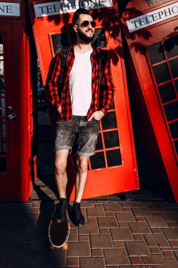 sakallı bir adam, güneş gözlüklü bir hipster, kırmızı bir telefon kulübesinin önünde kaykayla poz verdi.