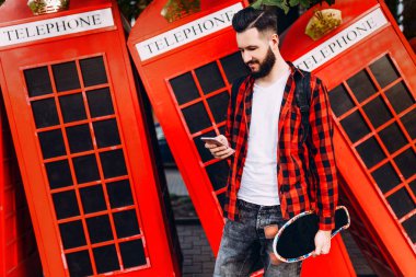 Sakallı genç adam, kaykaylı ekose gömlek giymiş hipster, elinde bir akıllı telefon tutuyor