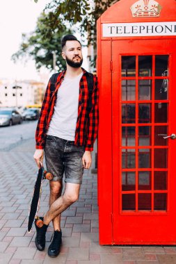 Sakallı şık adam, elinde kaykay tutan ve şehirde kırmızı bir telefon kulübesinin önünde poz veren bir adam.