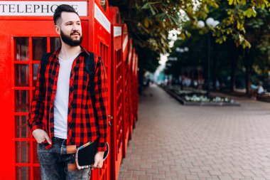 Sakallı şık adam, elinde kaykay tutan ve şehirde kırmızı bir telefon kulübesinin önünde poz veren bir adam.