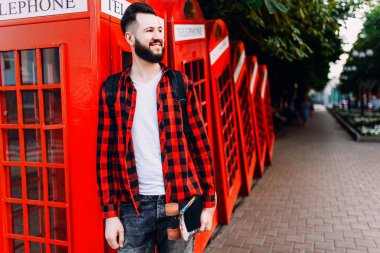 Sakallı şık adam, elinde kaykay tutan ve şehirde kırmızı bir telefon kulübesinin önünde poz veren bir adam.