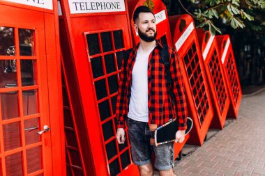 Sakallı şık adam, elinde kaykay tutan ve şehirde kırmızı bir telefon kulübesinin önünde poz veren bir adam.