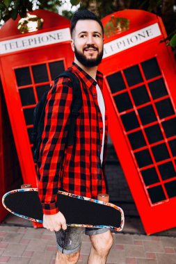 Sakallı şık adam, elinde kaykay tutan ve şehirde kırmızı bir telefon kulübesinin önünde poz veren bir adam.