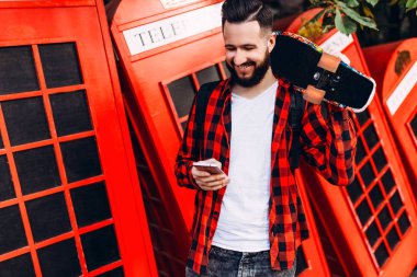 Sakallı genç adam, kaykaylı ekose gömlek giymiş hipster, elinde bir akıllı telefon tutuyor
