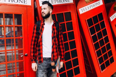 Sakallı şık adam, elinde kaykay tutan ve şehirde kırmızı bir telefon kulübesinin önünde poz veren bir adam.
