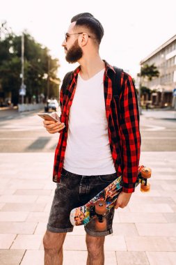 sakallı adam, elinde bir kaykay ve akıllı telefon ile şehirde dolaşıyor