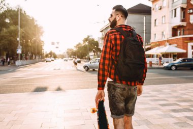 Kablosuz kulaklık bir sakallı şık adam şehir etrafında bir kaykay ile yürüyor