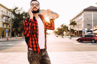 sakallı adam, bir kaykay ile şehir etrafında yürür ve bir akıllı telefon konuşuyor