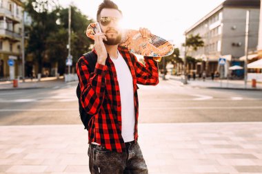 sakallı adam, bir kaykay ile şehir etrafında yürür ve bir akıllı telefon konuşuyor