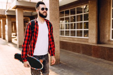 Kablosuz kulaklık bir sakallı şık adam şehir etrafında bir kaykay ile yürüyor