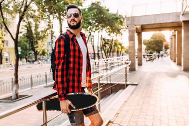 Kablosuz kulaklık bir sakallı şık adam şehir etrafında bir kaykay ile yürüyor