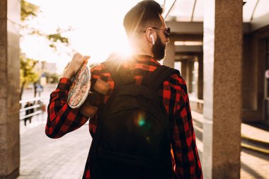 Kablosuz kulaklık bir sakallı şık adam şehir etrafında bir kaykay ile yürüyor