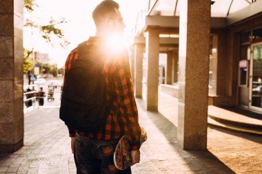 Kablosuz kulaklık sakallı şık adam bir kaykay ile şehir etrafında yürüyor