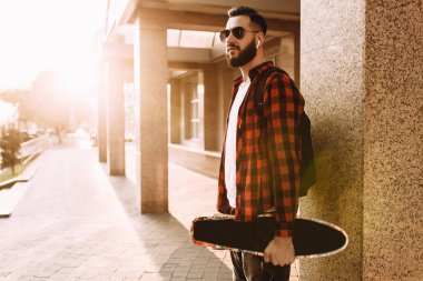 Kablosuz kulaklık sakallı şık adam bir kaykay ile şehir etrafında yürüyor