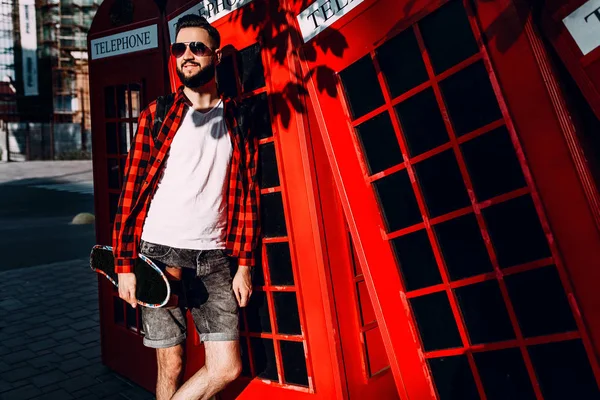 Homem elegante com barba, hipster segura um skate em suas mãos e posa na frente de uma cabine telefônica vermelha — Fotografia de Stock