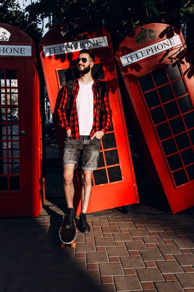 En man med skägg, en Hipster i solglasögon, poserar med en skateboard framför en röd telefon låda — Stockfoto