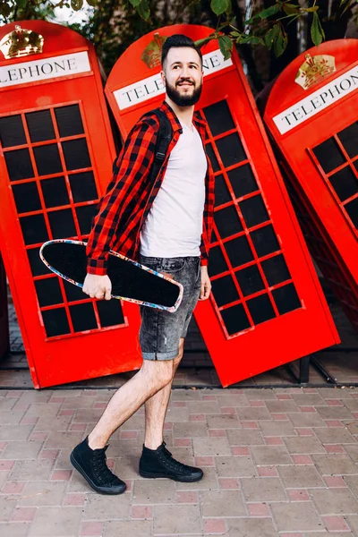 Snygg man med skägg, en man som håller en skateboard i händerna och poserar i staden framför en röd telefon låda — Stockfoto
