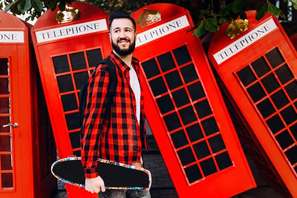 Snygg man med skägg, en man som håller en skateboard i händerna och poserar i staden framför en röd telefon låda — Stockfoto