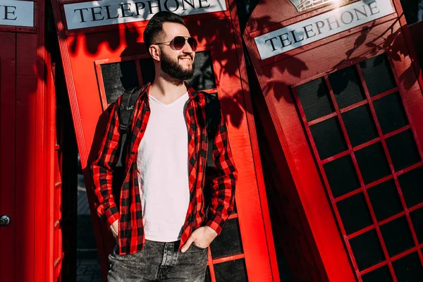 Hipster med skägg, en man i en vit T-shirt och solglasögon poserar i staden mittemot den röda telefon boxen — Stockfoto