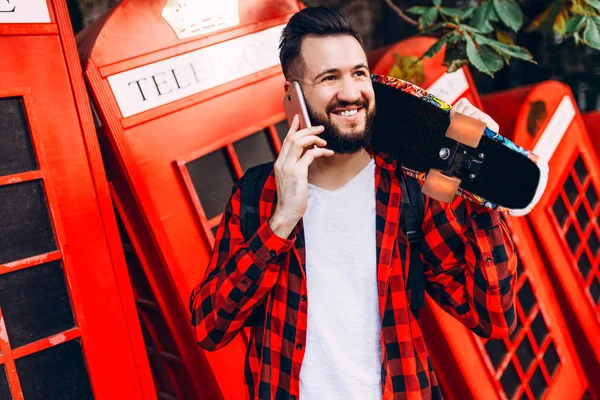 Man med skägg, hipster klädd i en pläd skjorta med en skateboard, pratar på en mobiltelefon — Stockfoto