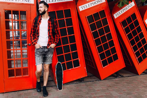 Чоловік з бородою, хіпстер в сонцезахисних окулярах, позує зі скейтбордом перед червоною телефонною коробкою — стокове фото