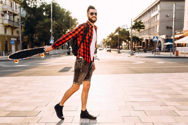 Ragazzo con la barba in occhiali da sole, cammina con uno skateboard al tramonto . — Foto Stock