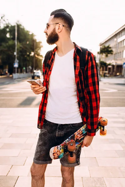 sakallı adam, elinde bir kaykay ve akıllı telefon ile şehirde dolaşıyor