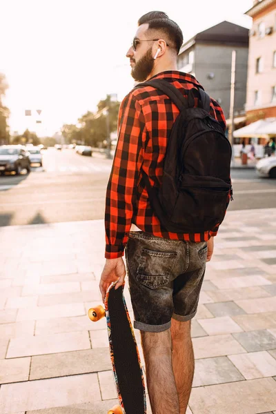 Kablosuz kulaklık bir sakallı şık adam şehir etrafında bir kaykay ile yürüyor