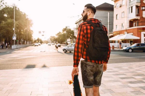 Kablosuz kulaklık bir sakallı şık adam şehir etrafında bir kaykay ile yürüyor