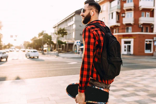 Kablosuz kulaklık bir sakallı şık adam şehir etrafında bir kaykay ile yürüyor