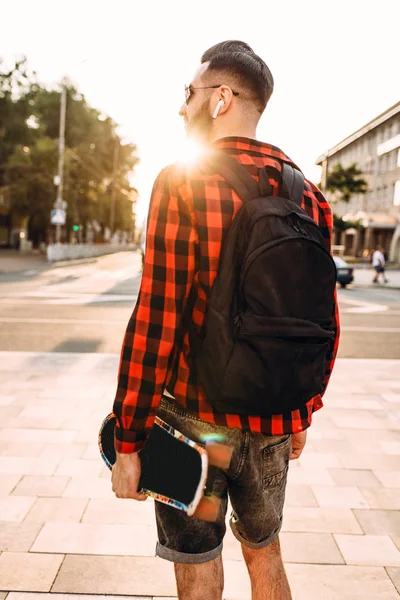 Kablosuz kulaklık bir sakallı şık adam şehir etrafında bir kaykay ile yürüyor