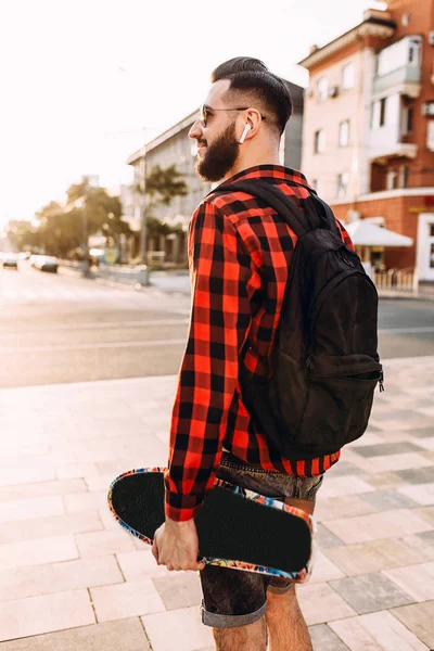 Elegante ragazzo con la barba in cuffie wireless passeggiate con uno skateboard in giro per la città — Foto Stock