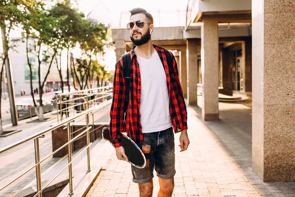 Stylish guy with a beard in wireless headphones walks with a skateboard around the city — Stock Photo, Image