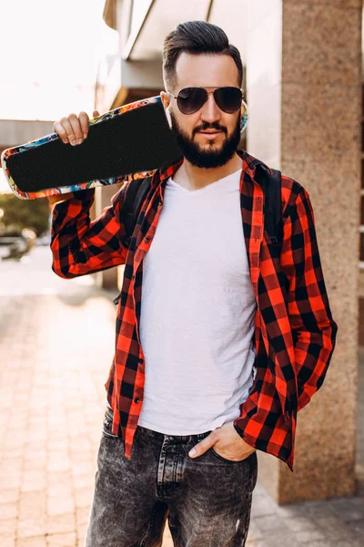 Стильний хлопець з бородою в бездротових навушниках ходить зі скейтбордом навколо міста — стокове фото
