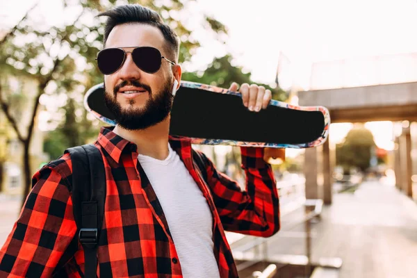 Un gars élégant avec une barbe dans un casque sans fil marche avec une planche à roulettes autour de la ville — Photo