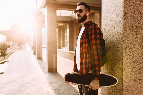 Kablosuz kulaklık sakallı şık adam bir kaykay ile şehir etrafında yürüyor