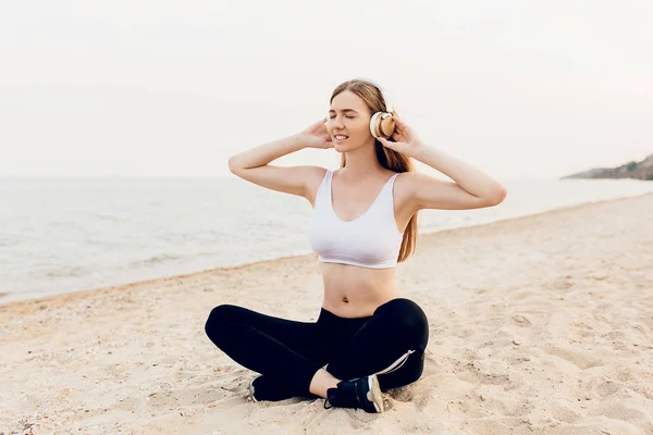 Sportief meisje zittend op het strand en luisteren naar muziek in headp — Stockfoto