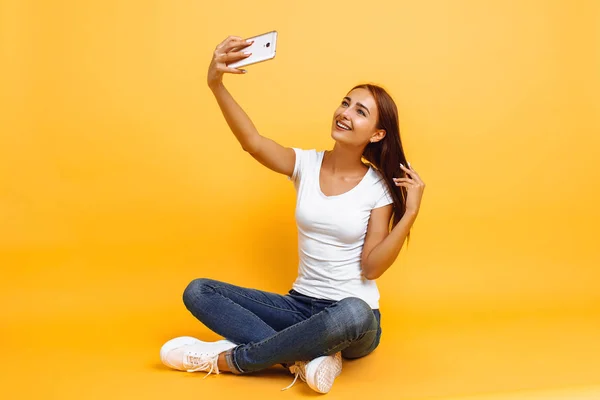 Chica joven en camiseta blanca sentada con las piernas cruzadas y tomando uno mismo — Foto de Stock