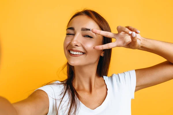 Fiatal lány veszi selfie és megmutatja a béke gesztus, a sárga backg — Stock Fotó