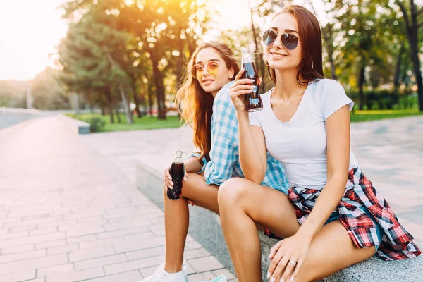 Две стильные девушки, выпьют напиток сидя в парке — стоковое фото