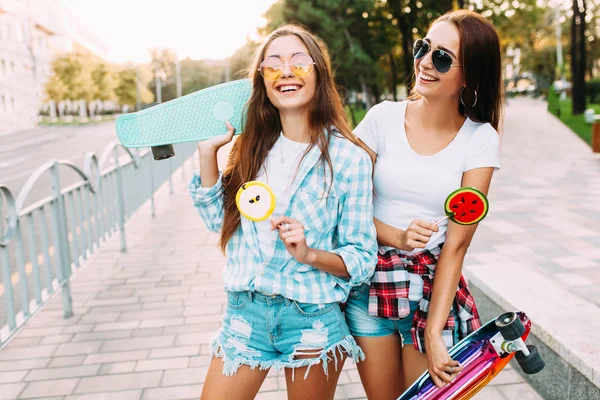 Due giovani ragazze alla moda passeggiano per la città soleggiata con lecca-lecca — Foto Stock