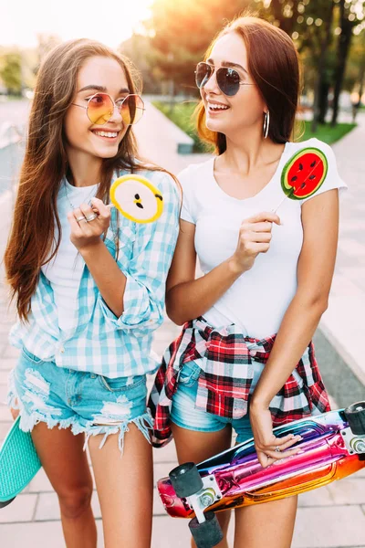 Dos chicas jóvenes y elegantes caminan por la ciudad soleada con piruleta —  Fotos de Stock
