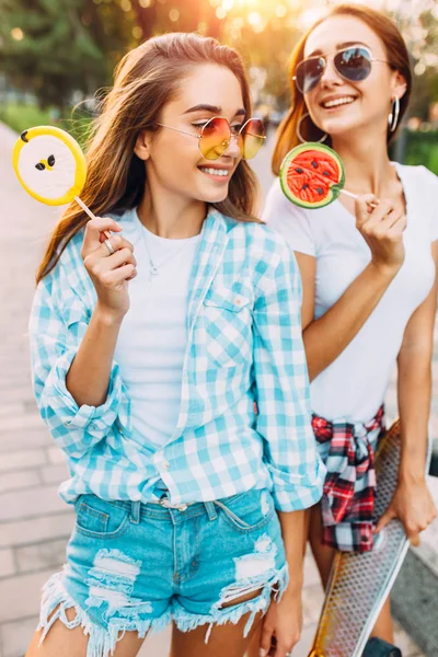 Due giovani ragazze alla moda passeggiano per la città soleggiata con lecca-lecca — Foto Stock