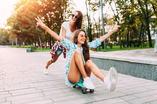 Две стильные взволнованные девушки развлекаются и катаются на скейтборде в парке — стоковое фото