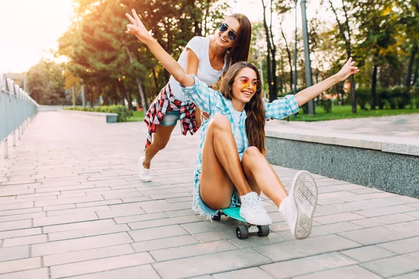 Duas meninas animadas elegantes se divertir e skate no parque em — Fotografia de Stock