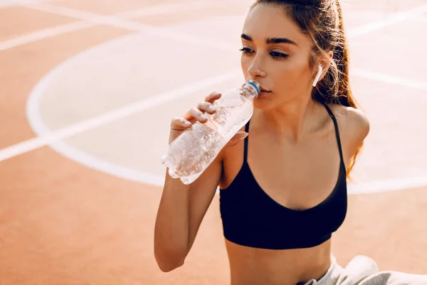 Sportliches Mädchen, in Top und Jogginghose, trinkt Wasser aus der Flasche, sitzt nach dem Training auf dem Feld — Stockfoto